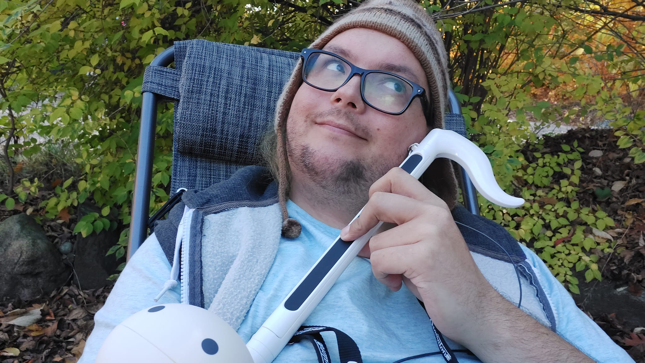 A man is wearing a toque, reclining in a lawn chair, and holding an otomatone, an electronic musical insturment shaped like a music note